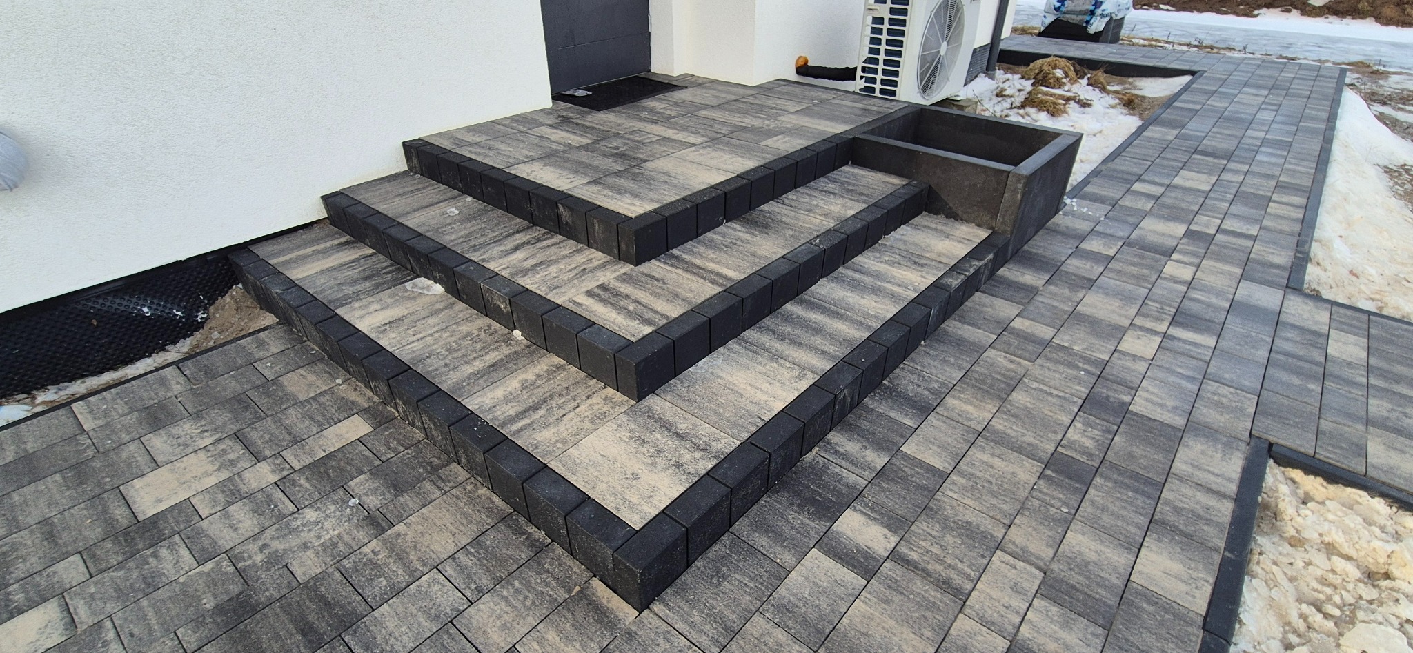Schody i chodnik z kostki brukowej w odcieniach szarości, z czarnym obramowaniem, prowadzące do białego budynku. Widoczny fragment klimatyzatora i resztki śniegu.