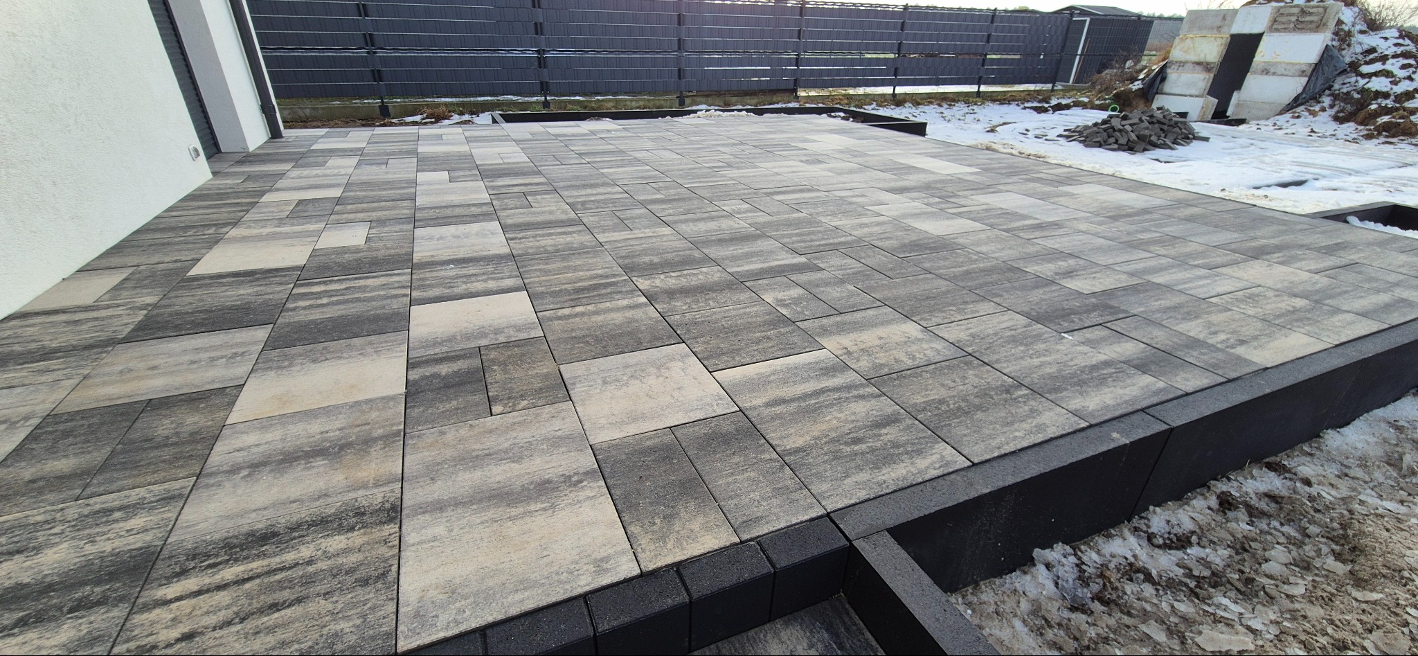 Ułożona kostka brukowa w Nakle nad Notecią. Szaro-beżowa kostka tworzy wzór na tarasie. Widoczne czarne obrzeża i fragmenty śniegu.