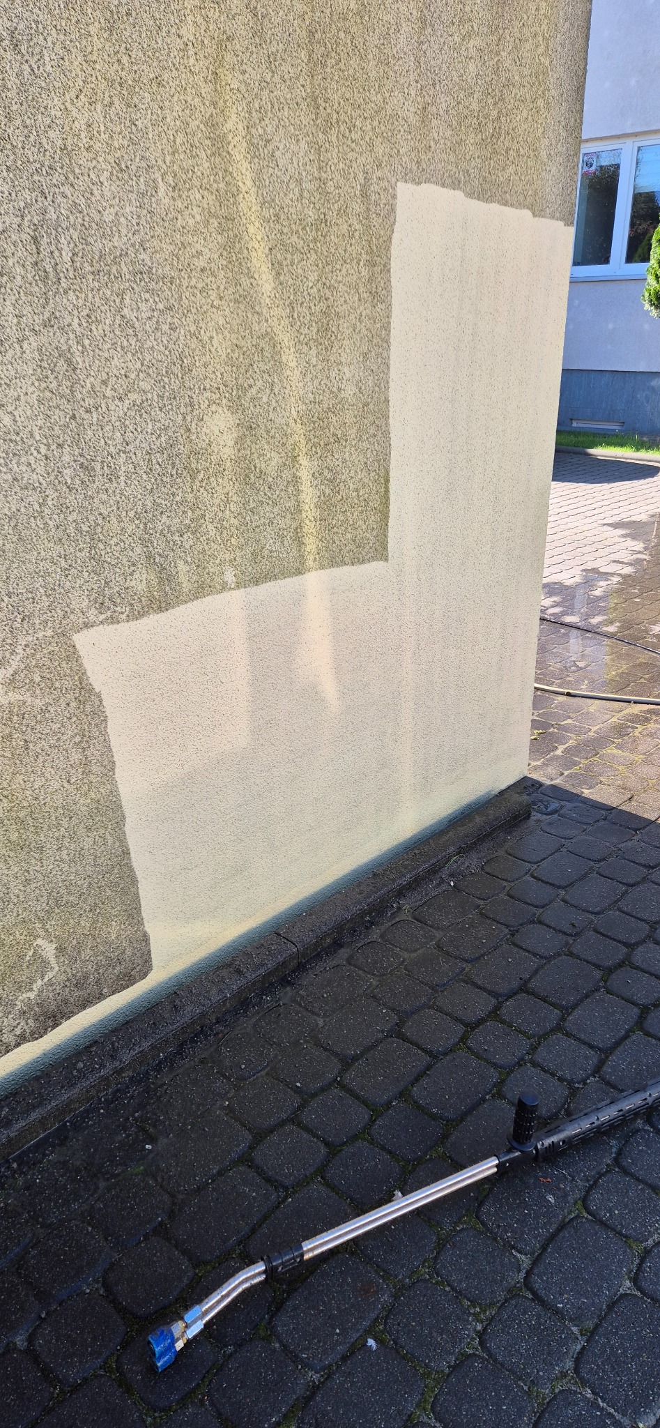 Fragment elewacji budynku z widocznym efektem czyszczenia. Część ściany jest czysta, a część pokryta zabrudzeniami. Na dole leży lanca do myjki ciśnieniowej.