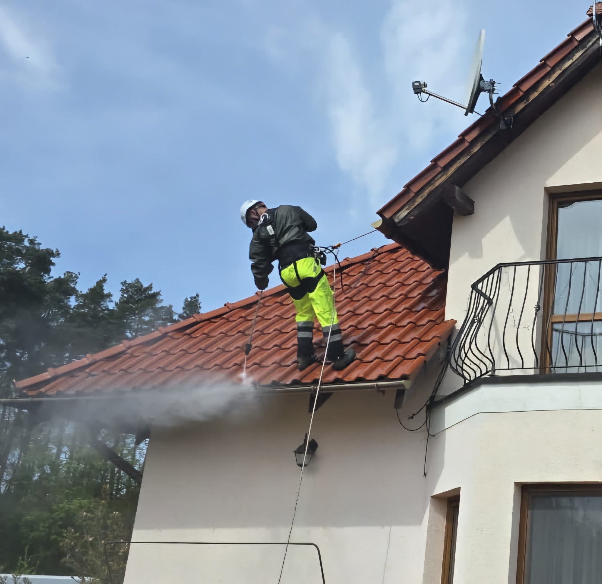 Pracownik w kasku i jaskrawych spodniach, zabezpieczony linami, czyści dach z czerwonej dachówki za pomocą myjki ciśnieniowej. Widoczny budynek z balkonem i anteną satelitarną.