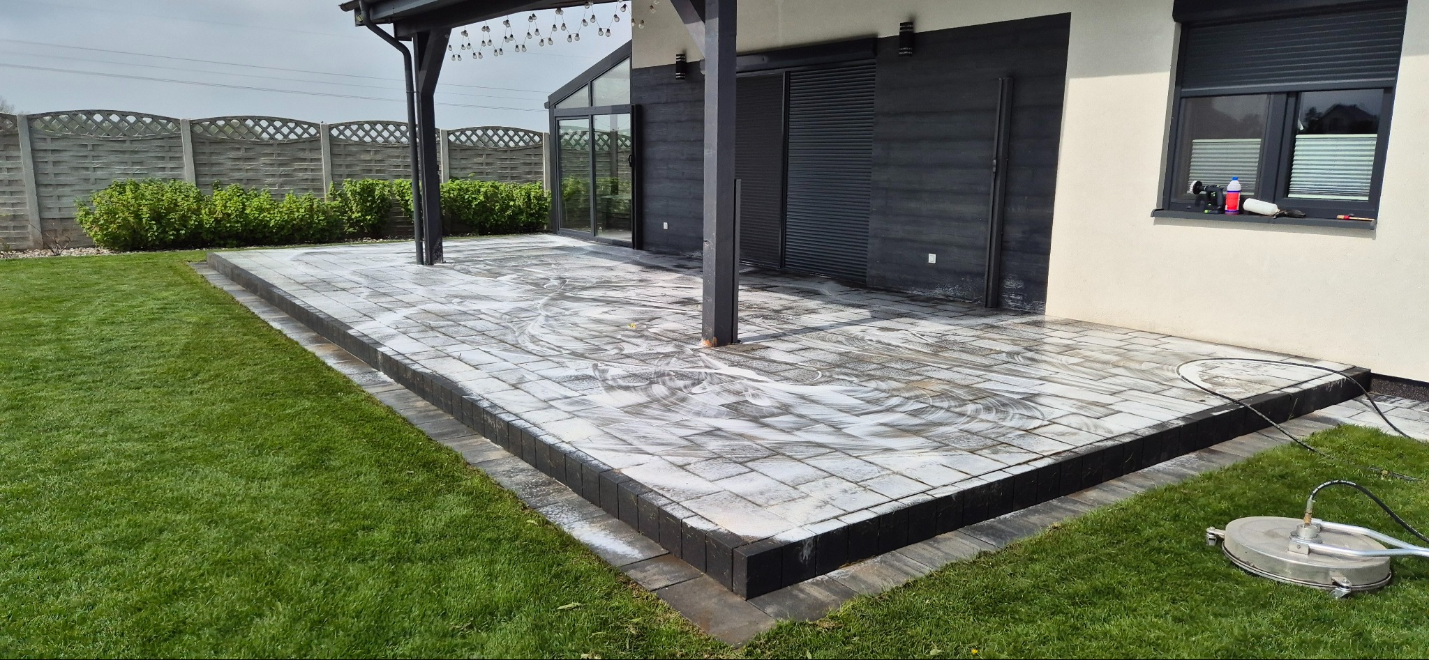 Kostka brukowa na tarasie w trakcie czyszczenia myjką ciśnieniową. Widoczne ślady piany i czyszczona powierzchnia. W tle dom z ciemną elewacją i zadbany trawnik.