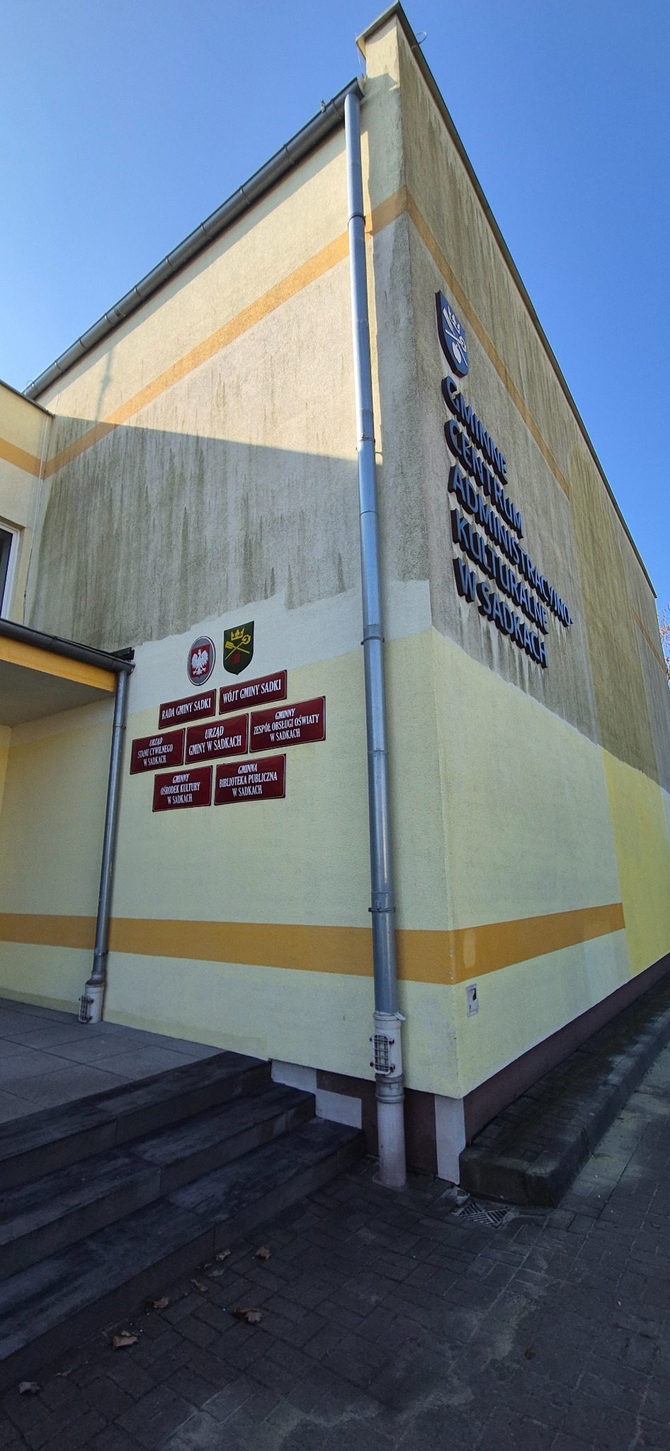 Żółta elewacja budynku Gminnego Centrum Administracyjno-Kulturalnego w Sadkach z widocznymi zabrudzeniami i zaciekami. Widoczne tabliczki informacyjne.