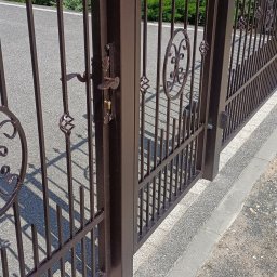 Mateusz Oleszek - Otwarta, brązowa brama wjazdowa z metalowymi zdobieniami, widoczny zamek i klamka, na tle asfaltowej drogi i zieleni. Ujęcie z bliska, z perspektywy przechodnia.