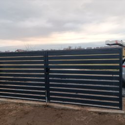 Mateusz Oleszek - Nowoczesna, pozioma brama wjazdowa w kolorze grafitowym, z widocznym mechanizmem otwierania i fragmentem ogrodzenia panelowego w tle. Pochmurne niebo.