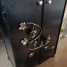 Mateusz Oleszek - Czarna, metalowa szafka z kutymi zdobieniami w kształcie winogron i liści, złote gałki, widok z boku, warsztatowe tło.