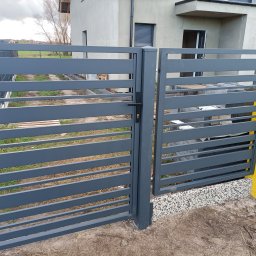 Mateusz Oleszek - Nowoczesna, pozioma brama wjazdowa w kolorze grafitowym, z czarną klamką, na tle jasnego domu jednorodzinnego i trawnika. Minimalistyczny design.