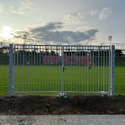 Mateusz Oleszek - Metalowa brama wjazdowa na boisko sportowe z widocznymi piłkarzami w oddali, oświetlona popołudniowym słońcem. Widoczne słupy oświetleniowe i siatka ogrodzeniowa.