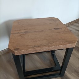 Mateusz Oleszek - Stolik kawowy z blatem imitującym drewno i czarną, metalową podstawą. Widoczne łączenia spawane. Ustawiony na drewnianej podłodze, na tle białej ściany.