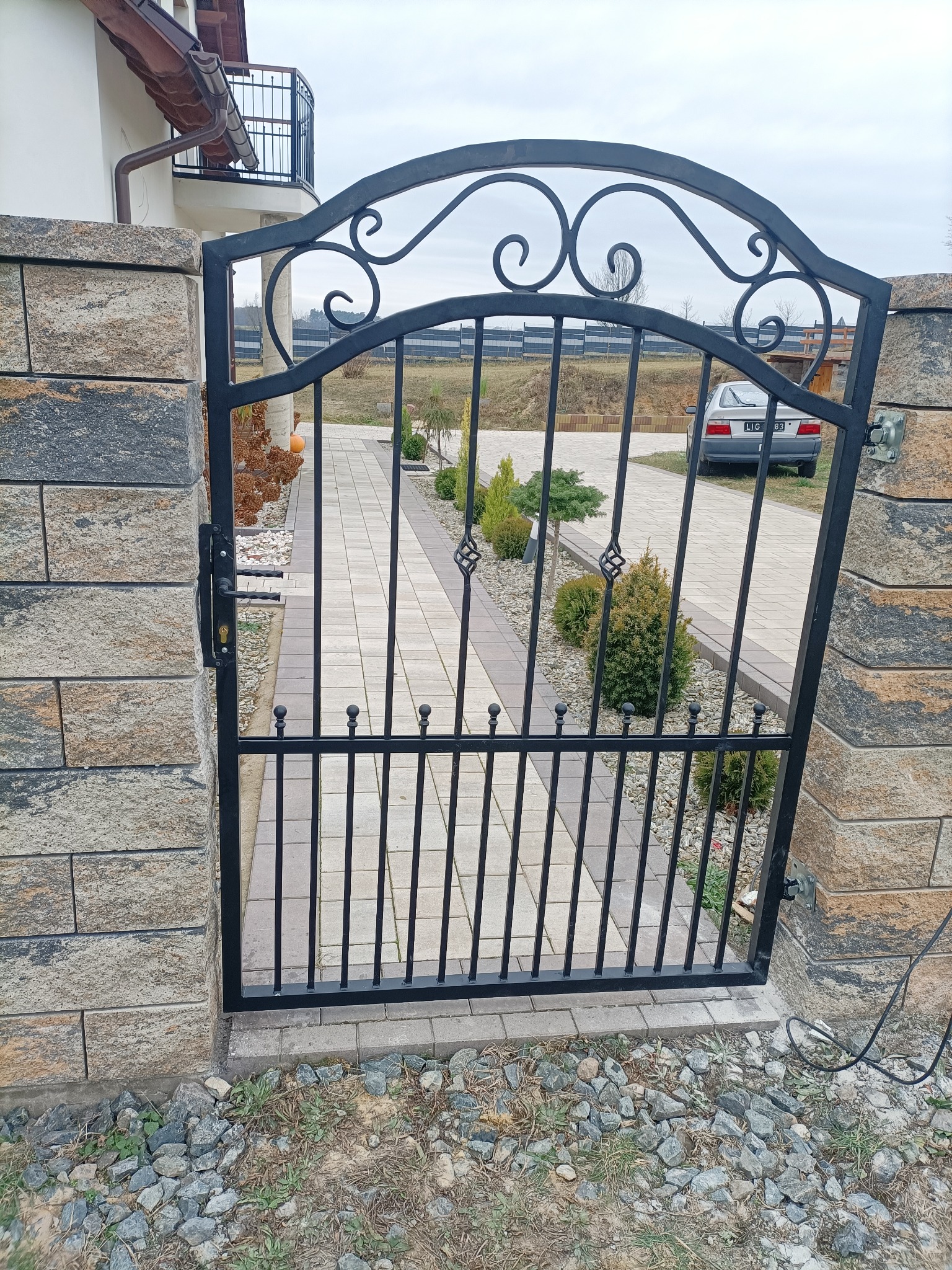 Czarna, metalowa furtka z ozdobnymi elementami, zamontowana między kamiennymi słupkami. Widok na ścieżkę i ogród. W tle zaparkowany samochód.