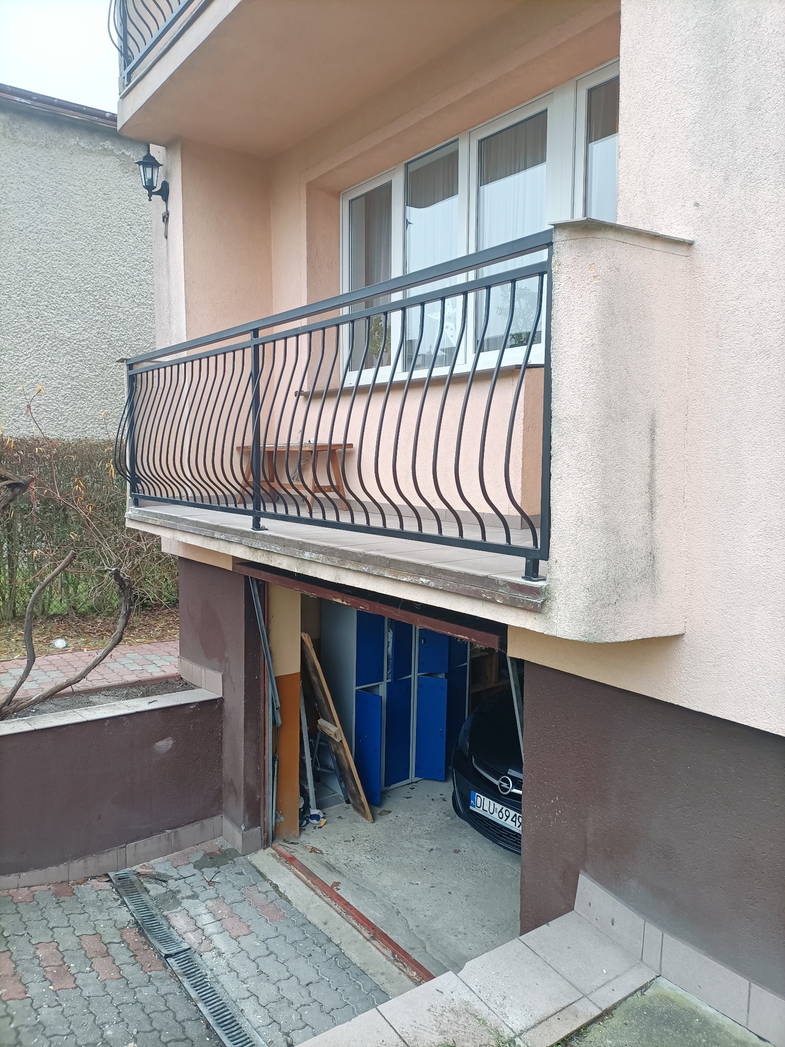 Balkon z czarną, metalową balustradą o falistym wzorze, widoczny z zewnątrz budynku. W tle okna i fragment wnętrza z meblami. Ujęcie z dołu, podkreślające detale balustrady.