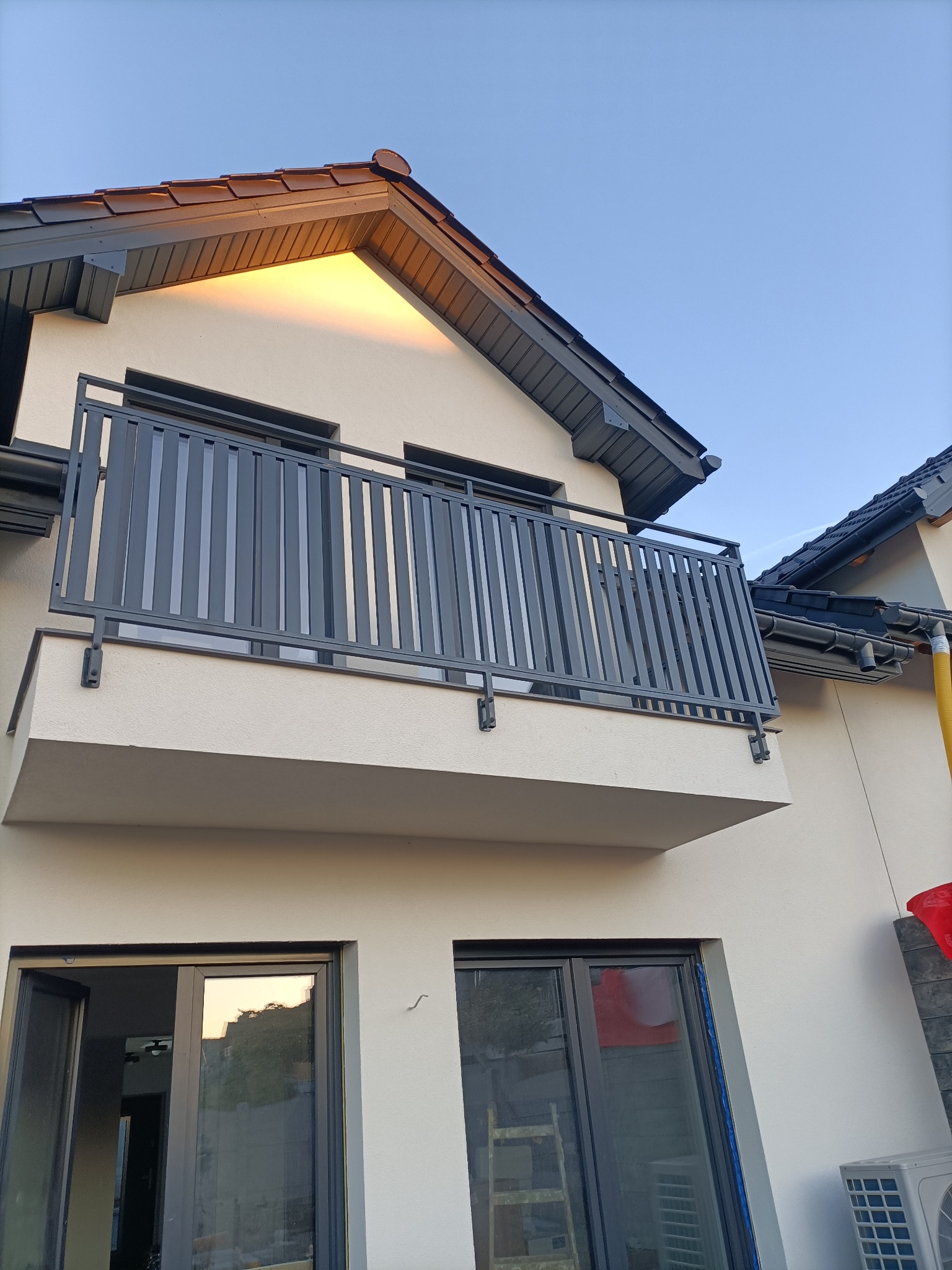 Szara, nowoczesna balustrada balkonowa z pionowych profili, zamontowana na jasnym budynku z ciemną dachówką. Widok z dołu, okna z odbiciami.