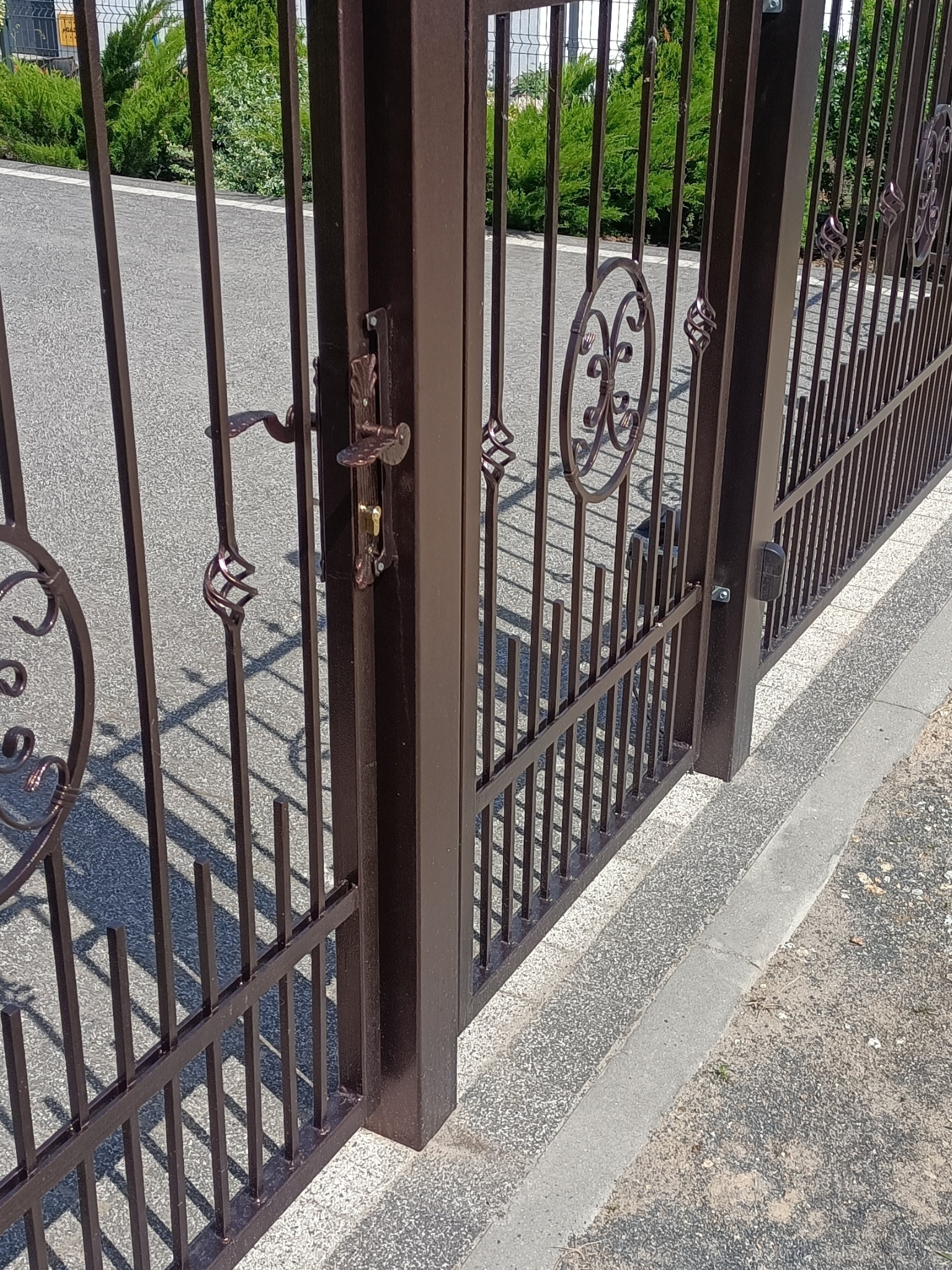 Otwarta, brązowa brama wjazdowa z metalowymi zdobieniami, widoczny zamek i klamka, na tle asfaltowej drogi i zieleni. Ujęcie z bliska, z perspektywy przechodnia.