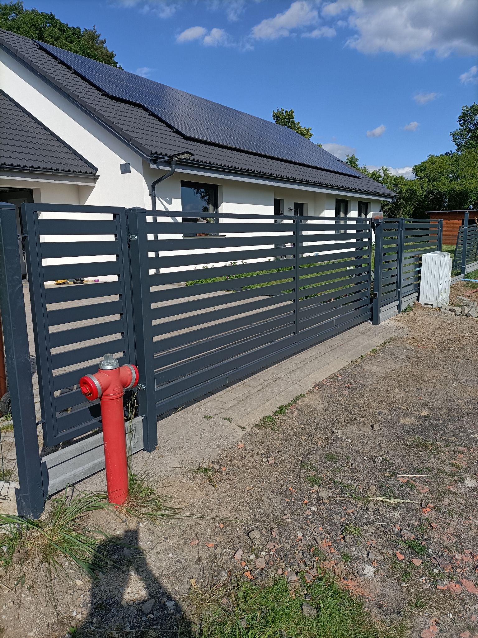 Nowoczesna, pozioma brama wjazdowa w kolorze grafitowym, z widocznym hydrantem w tle, na tle domu z panelami słonecznymi. Brama z poziomych profili.