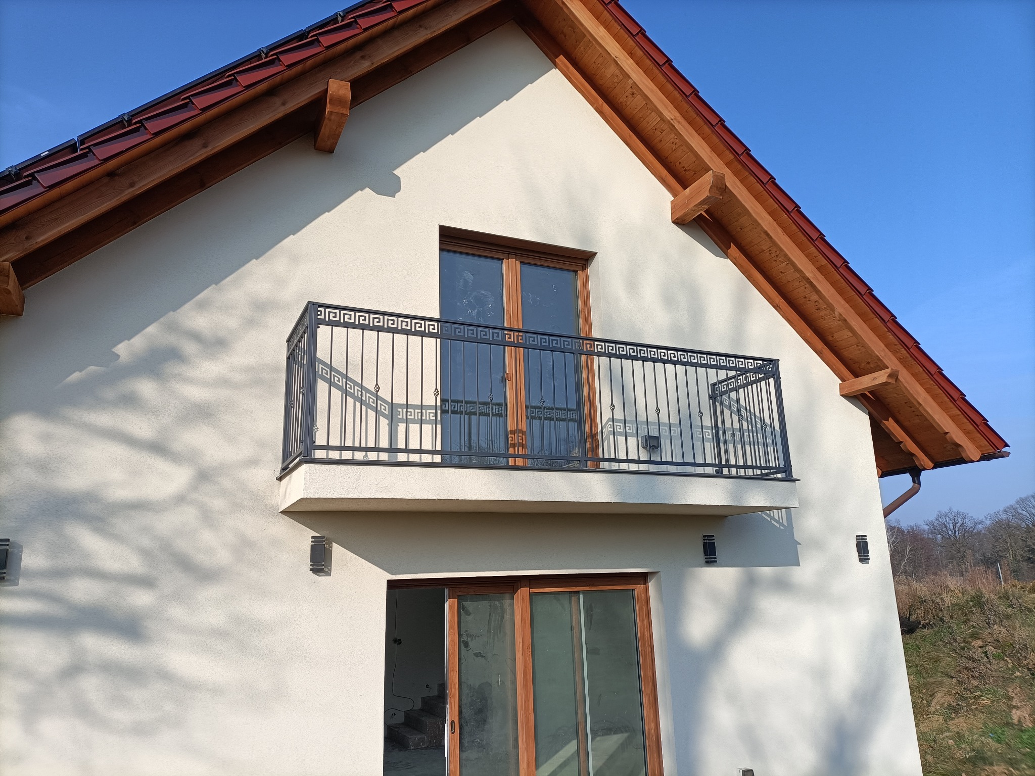 Elegancka, metalowa balustrada balkonowa z motywem greckim, zamontowana na jasnej elewacji domu z drewnianymi elementami dachu i oknami. Widok z dołu, błękitne niebo w tle.