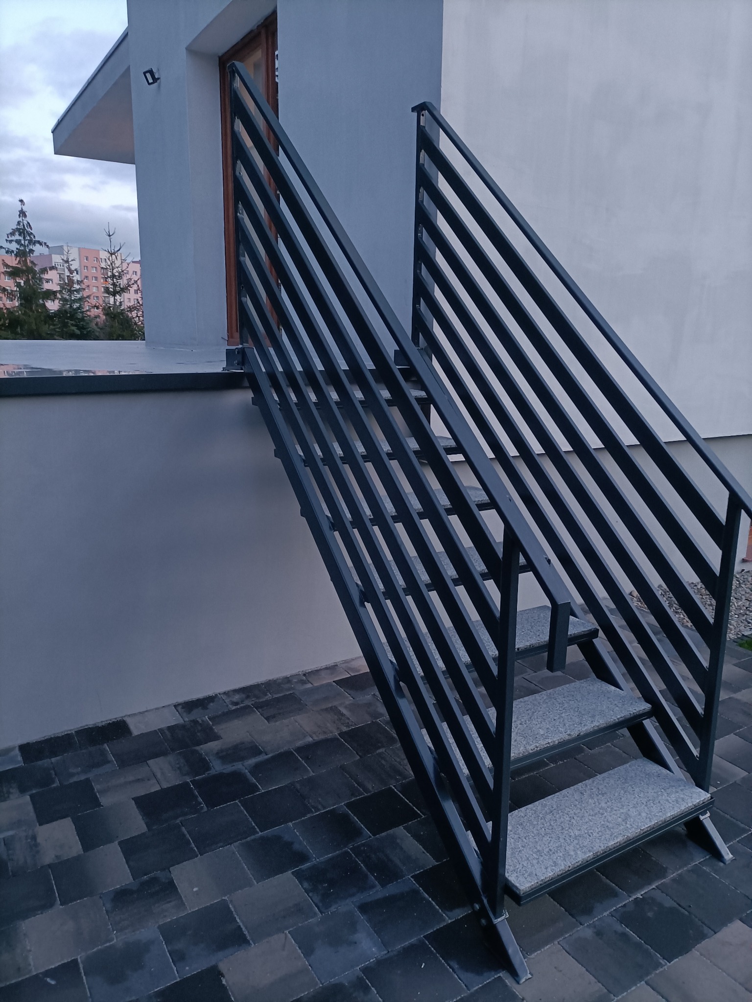 Nowoczesne, metalowe schody zewnętrzne z poręczą o minimalistycznym designie, prowadzące do wejścia budynku. Szare płytki na stopniach i ciemna kostka brukowa na podłożu.