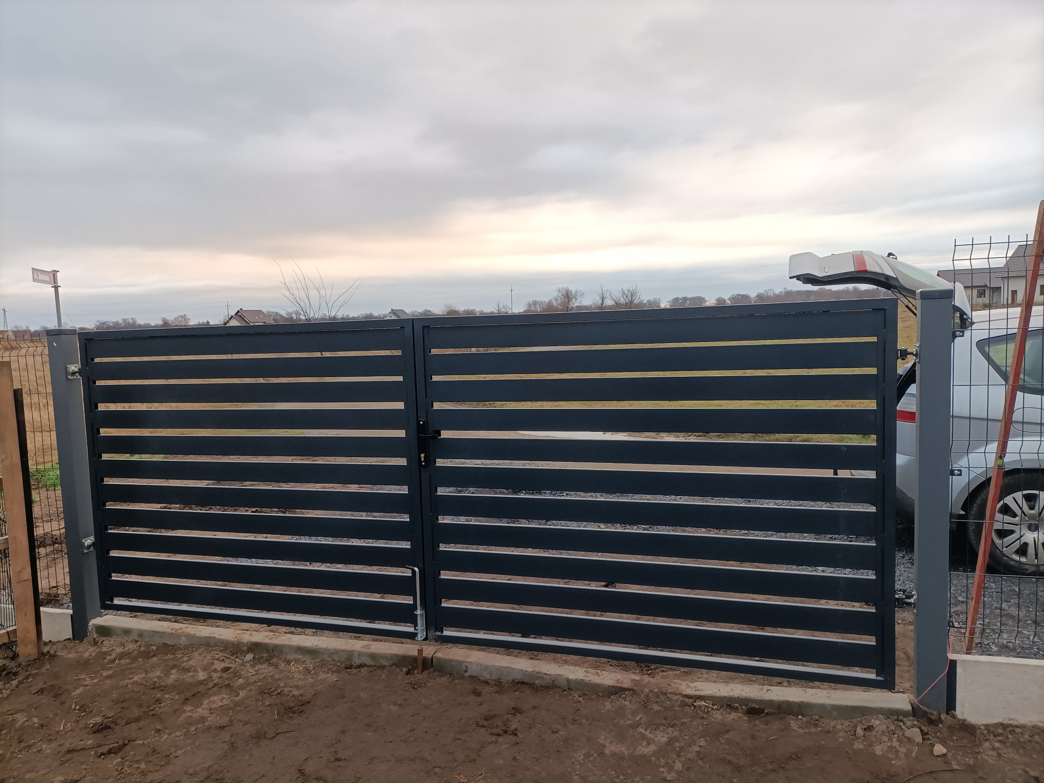 Nowoczesna, pozioma brama wjazdowa w kolorze grafitowym, z widocznym mechanizmem otwierania i fragmentem ogrodzenia panelowego w tle. Pochmurne niebo.