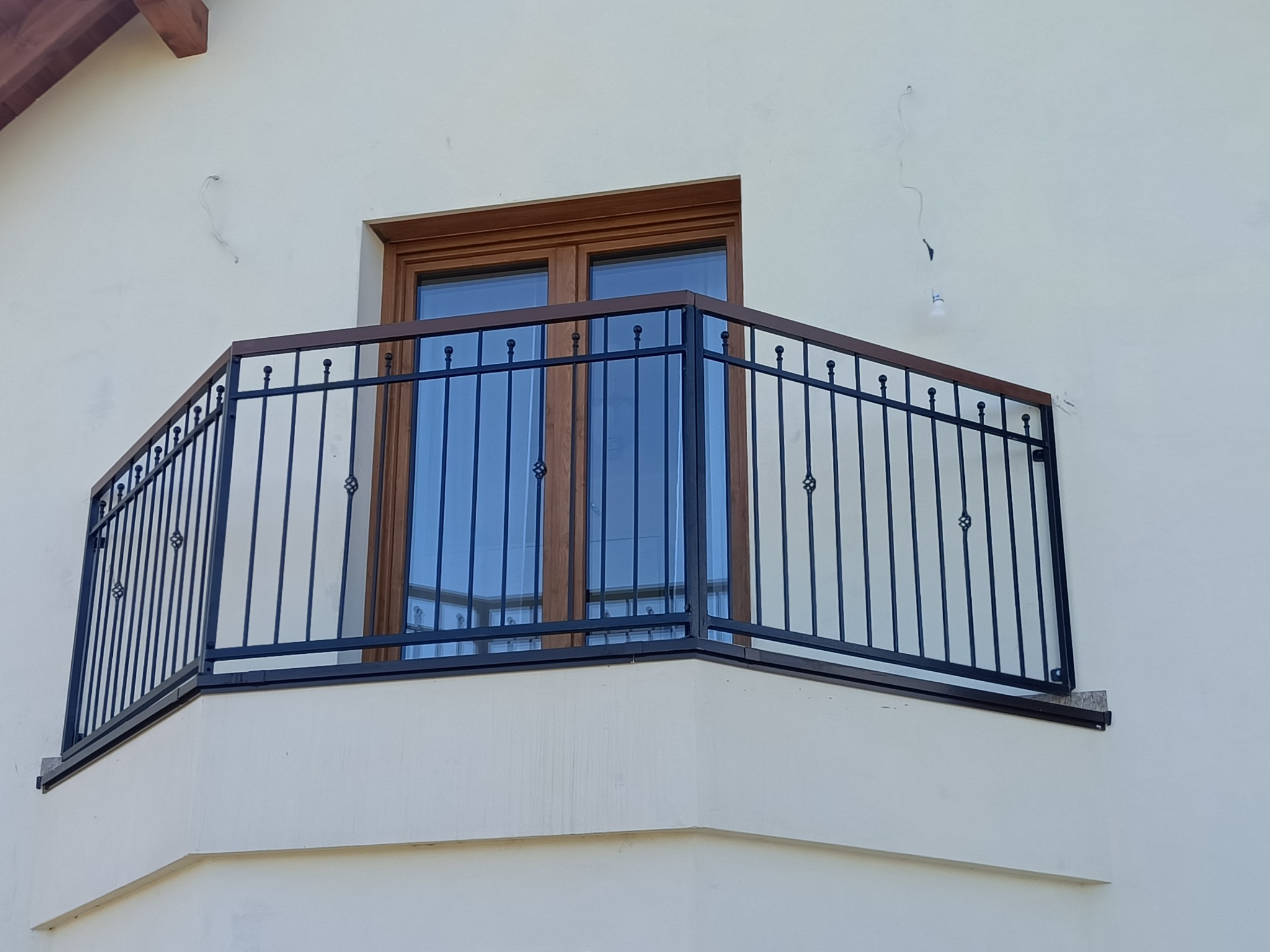 Kuta, czarna balustrada balkonowa z drewnianym wykończeniem, zamontowana na balkonie z beżową elewacją. Widoczne okno balkonowe z drewnianą ramą.