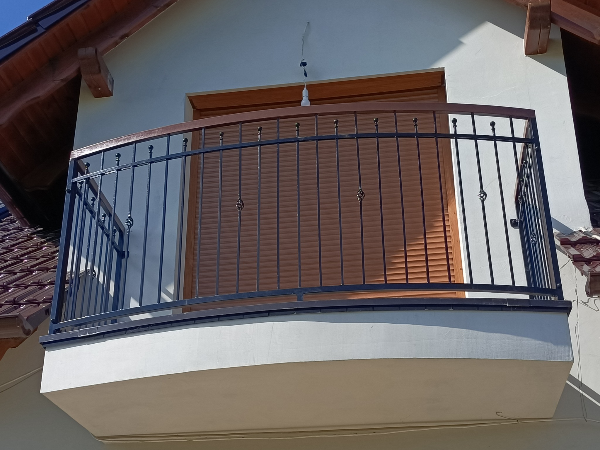 Kuta balustrada balkonowa z drewnianym elementem górnym, czarne pionowe pręty z ozdobnymi detalami, na tle zamkniętych, brązowych rolet okiennych.