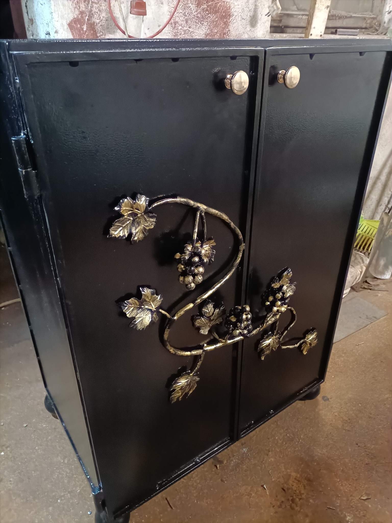 Czarna, metalowa szafka z kutymi zdobieniami w kształcie winogron i liści, złote gałki, widok z boku, warsztatowe tło.