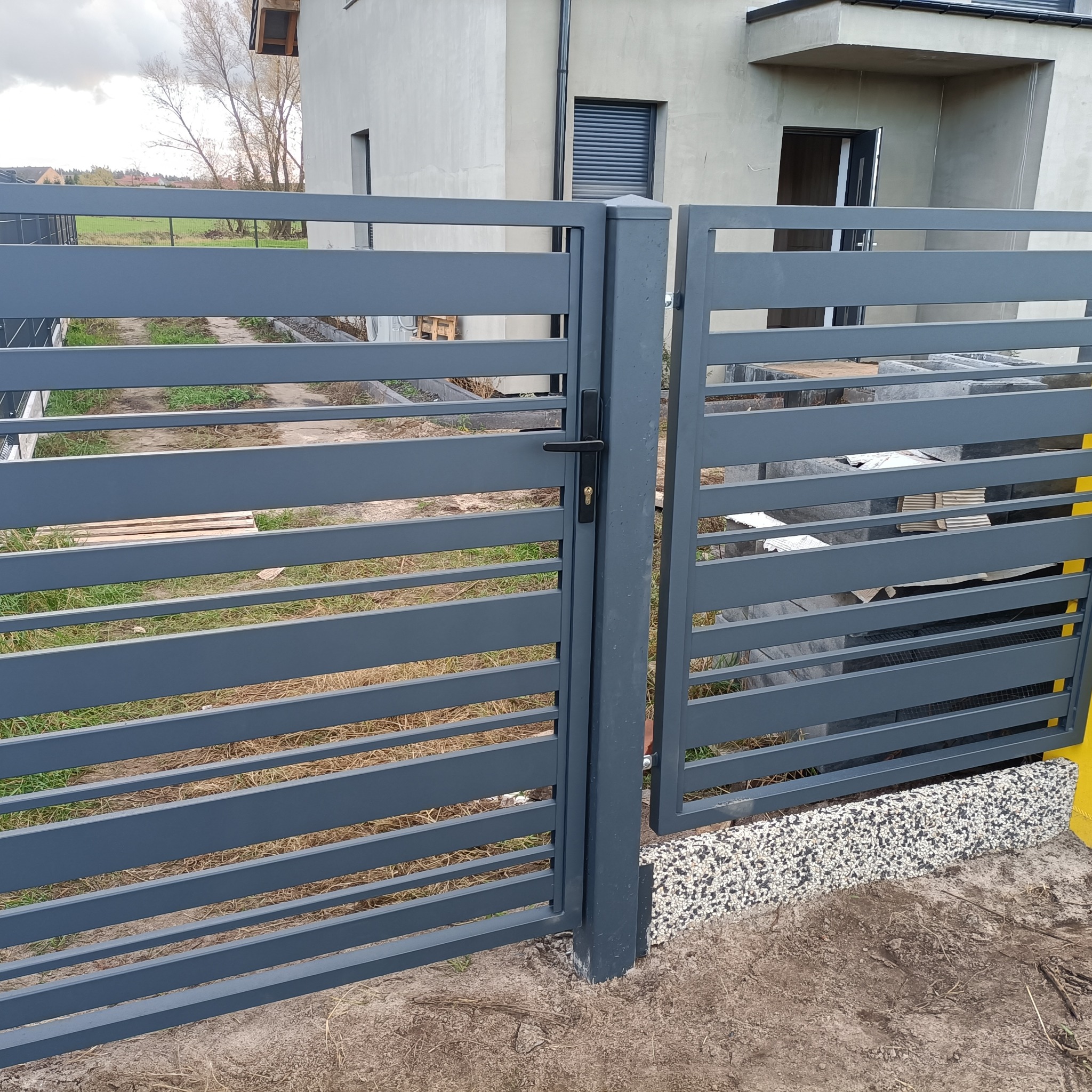 Nowoczesna, pozioma brama wjazdowa w kolorze grafitowym, z czarną klamką, na tle jasnego domu jednorodzinnego i trawnika. Minimalistyczny design.