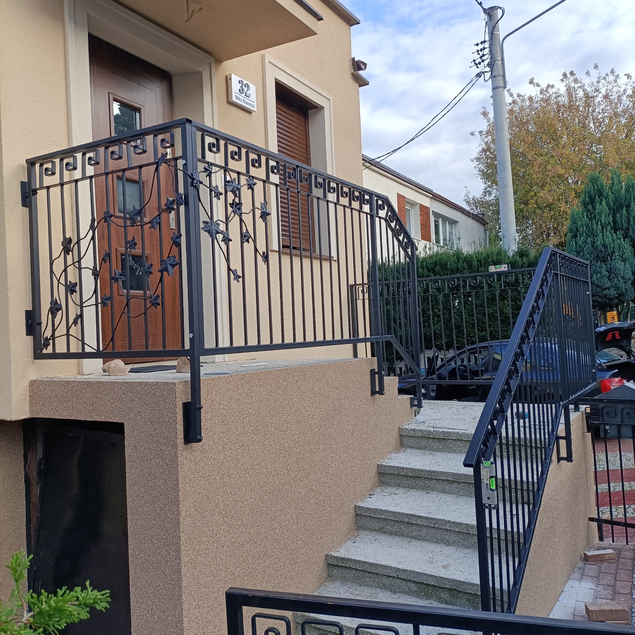 Kuta, czarna balustrada z motywem roślinnym zdobi wejście do domu z beżową elewacją. Widoczne schody wejściowe i fragment ulicy. Dekoracyjny detal architektoniczny.