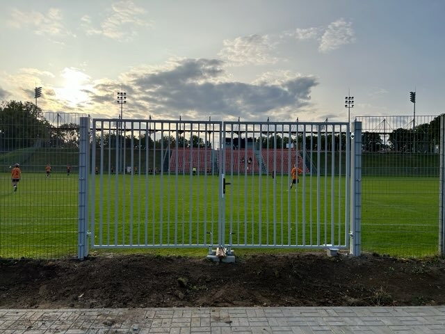 Metalowa brama wjazdowa na boisko sportowe z widocznymi piłkarzami w oddali, oświetlona popołudniowym słońcem. Widoczne słupy oświetleniowe i siatka ogrodzeniowa.