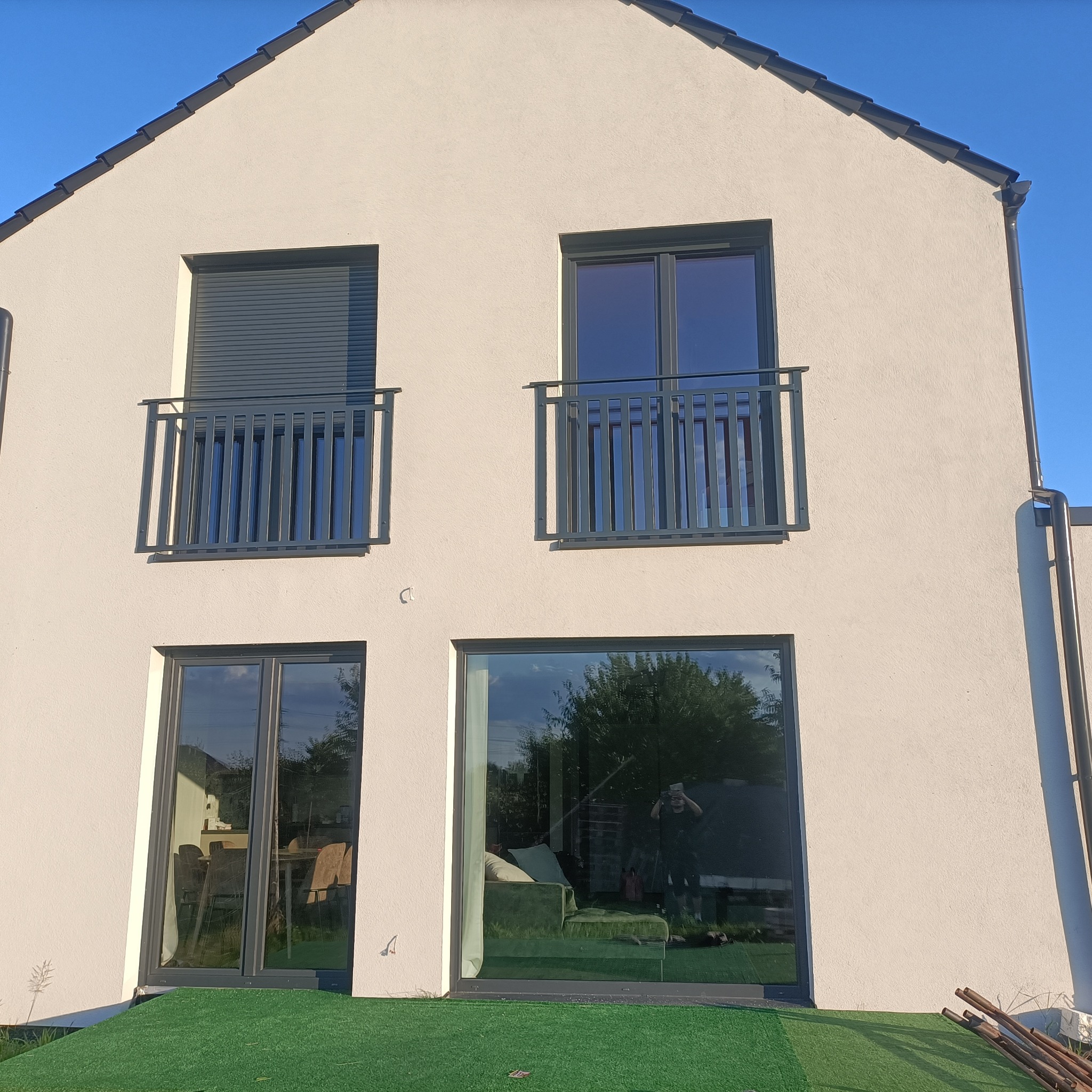 Nowoczesny dom z szarymi balustradami balkonowymi i dużymi oknami. Na dole sztuczna trawa. W oknach odbija się osoba robiąca zdjęcie.