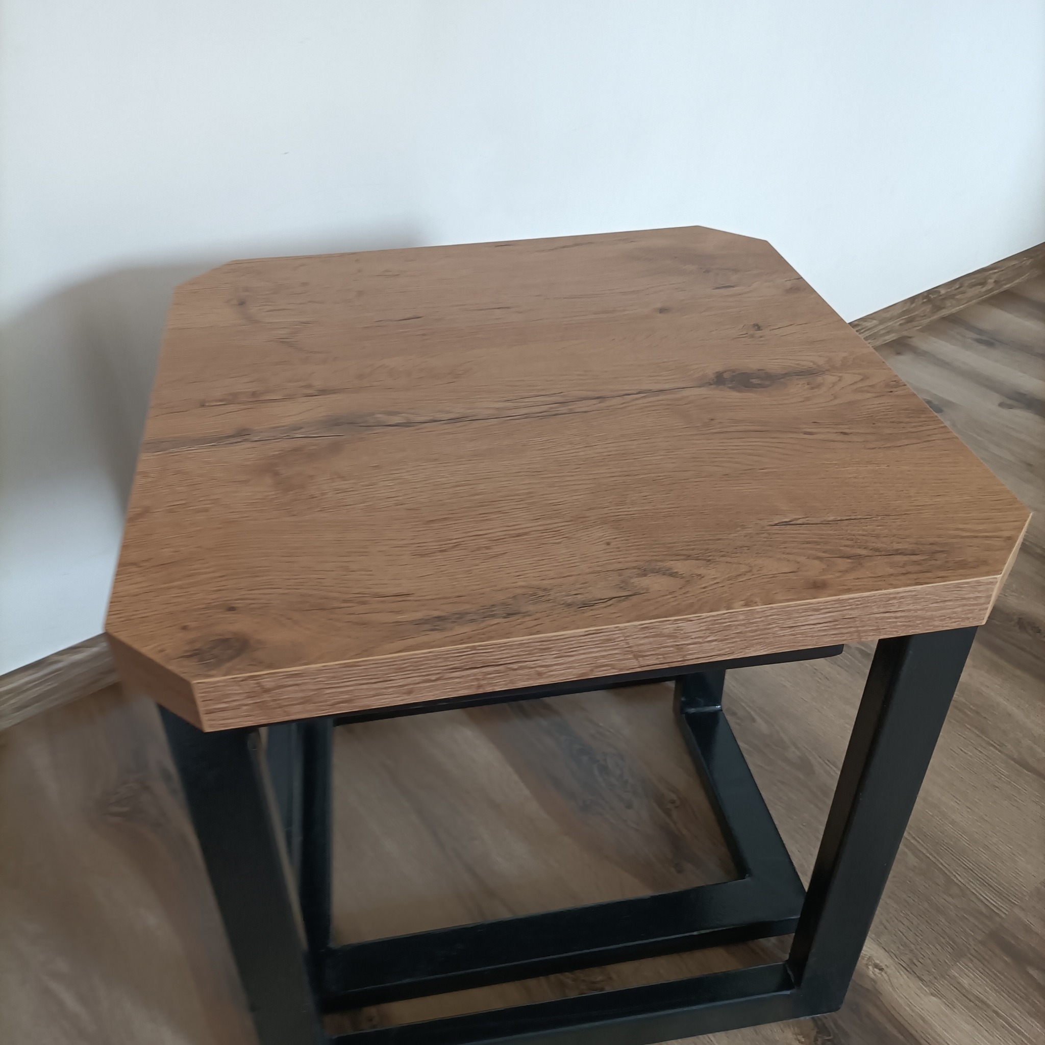 Stolik kawowy z blatem imitującym drewno i czarną, metalową podstawą. Widoczne łączenia spawane. Ustawiony na drewnianej podłodze, na tle białej ściany.