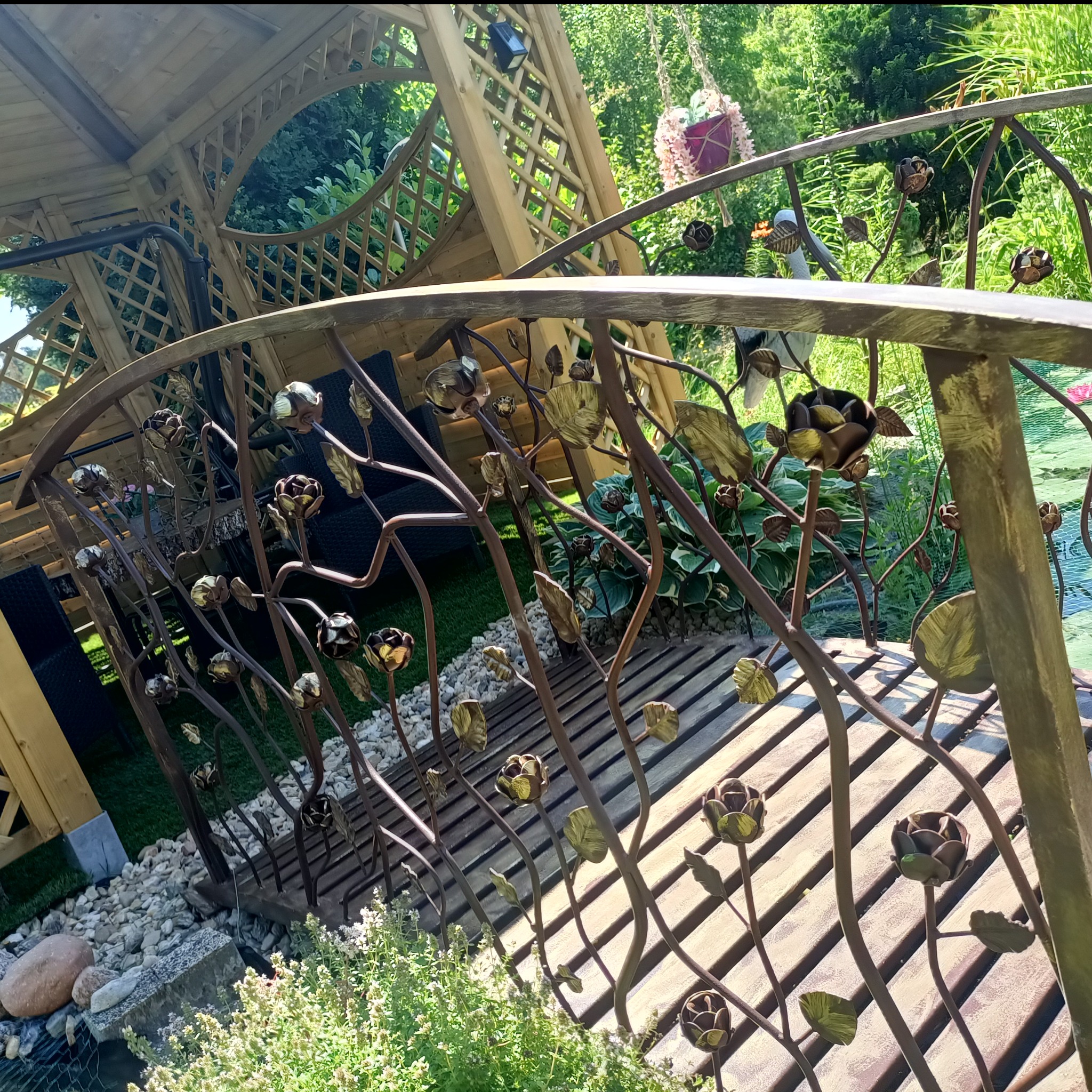 Dekoracyjny mostek ogrodowy z metalową balustradą w kształcie kwiatów, prowadzący do altany. Element małej architektury ogrodowej w słoneczny dzień.