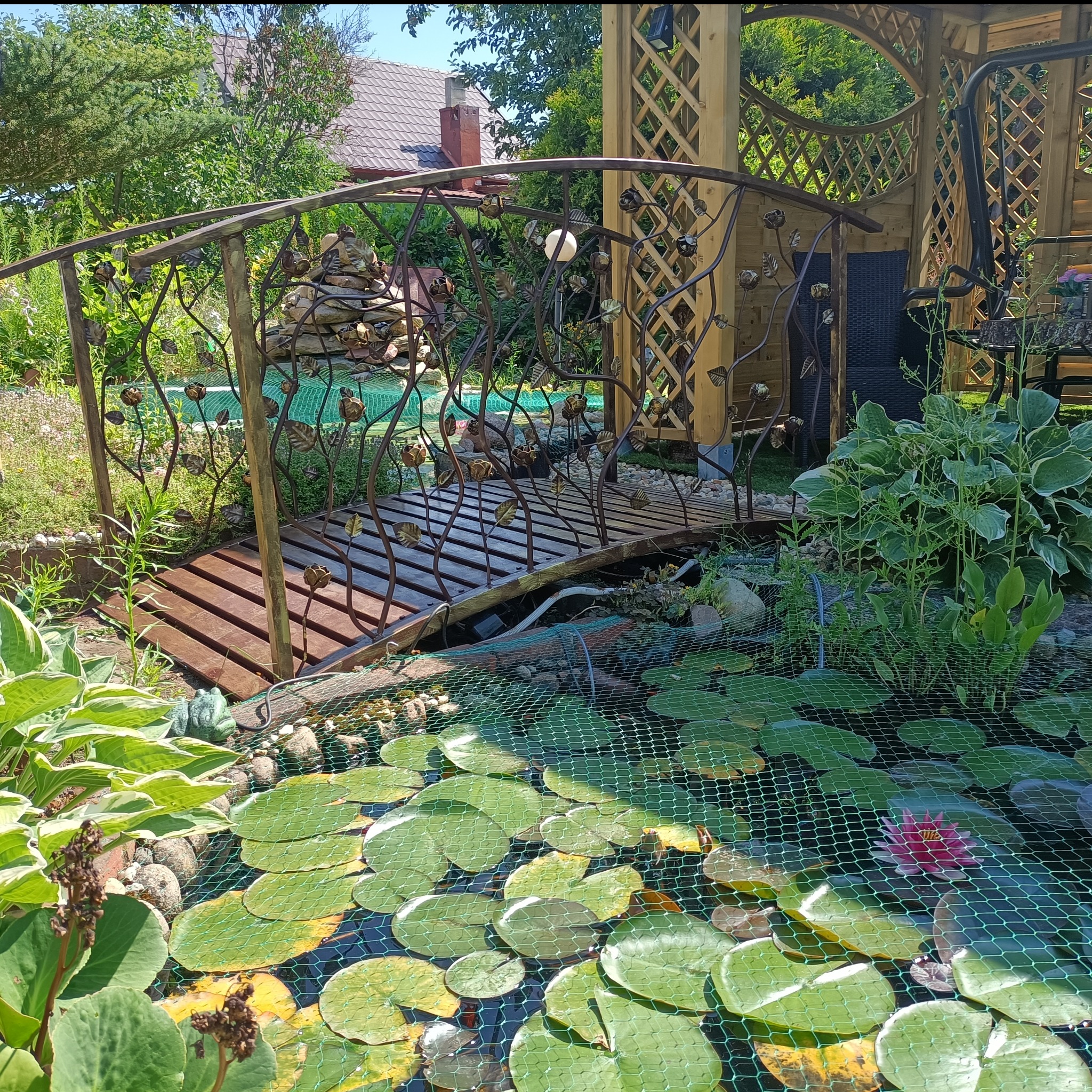 Kuty mostek nad oczkiem wodnym z liliami wodnymi, zdobiony motywem roślinnym. W tle altana z kratownicą i bujna zieleń ogrodu.