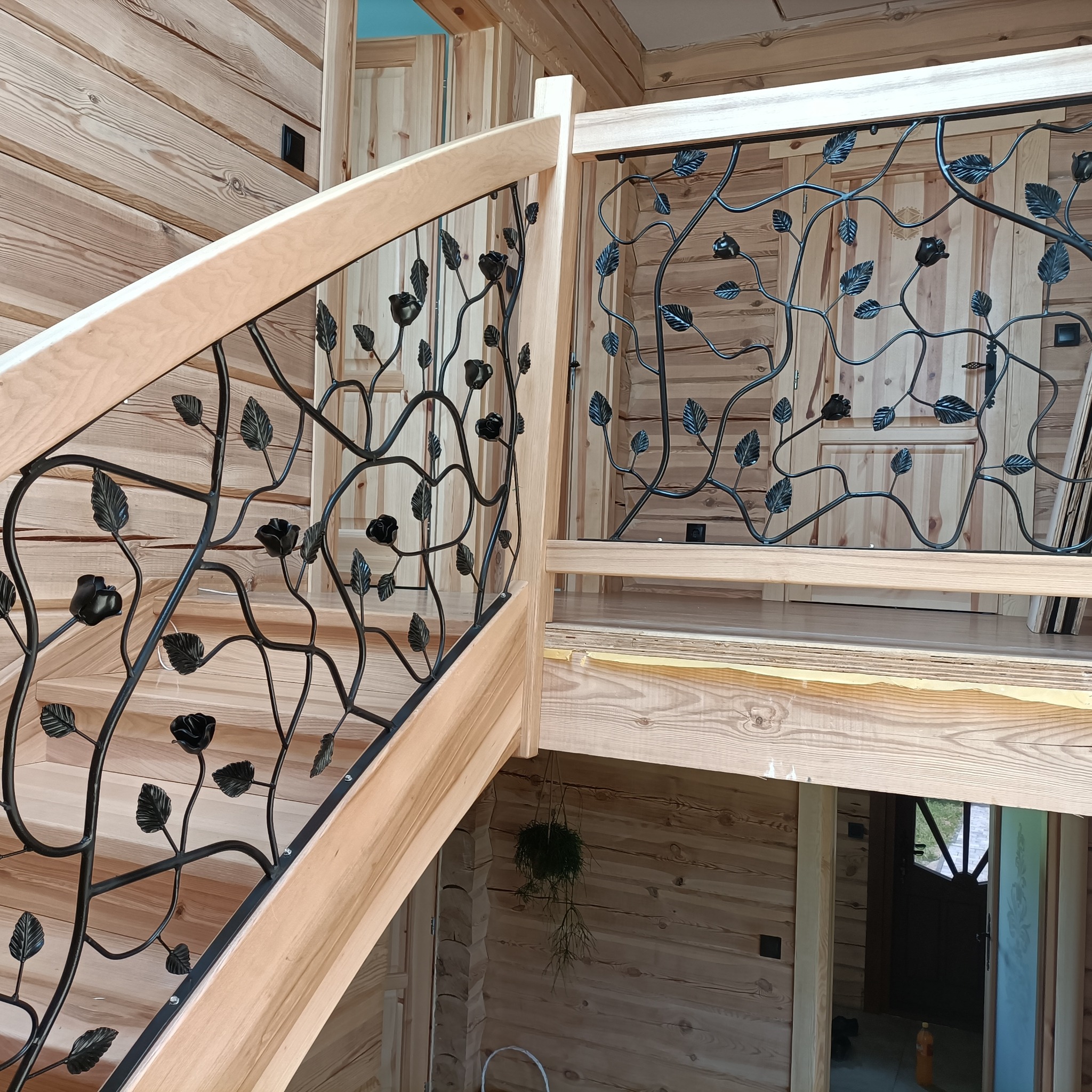 Kuta balustrada schodowa z motywem roślinnym, czarne metalowe elementy na jasnym drewnianym stelażu, wnętrze domu z bali, widok z dołu na górę.