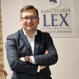 Lex Kancelaria Radcy Prawnego Wojciech Artemiuk - Uśmiechnięty prawnik w okularach i garniturze na tle logo Kancelarii Lex Wojciech Artemiuk, z zegarkiem na ręku, pozuje z założonymi rękami.