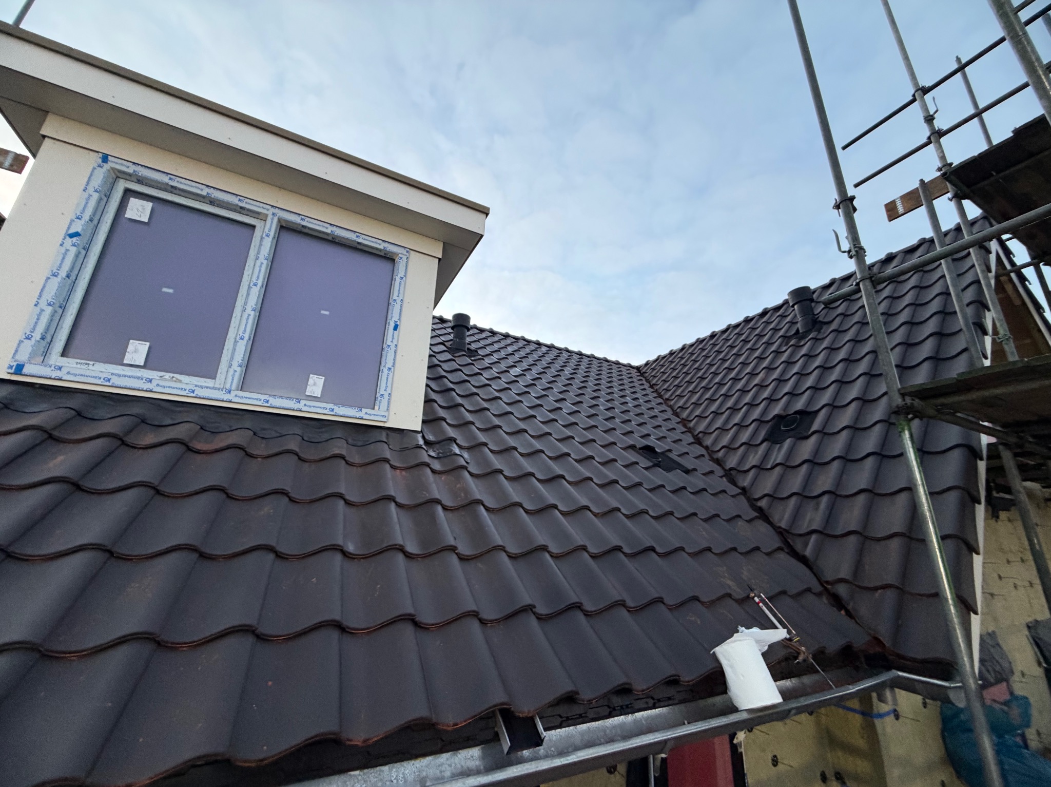 Ciemna dachówka ceramiczna na skośnym dachu z oknem połaciowym i rusztowaniem. Widok z dołu, podkreślający strukturę pokrycia i detale okna.