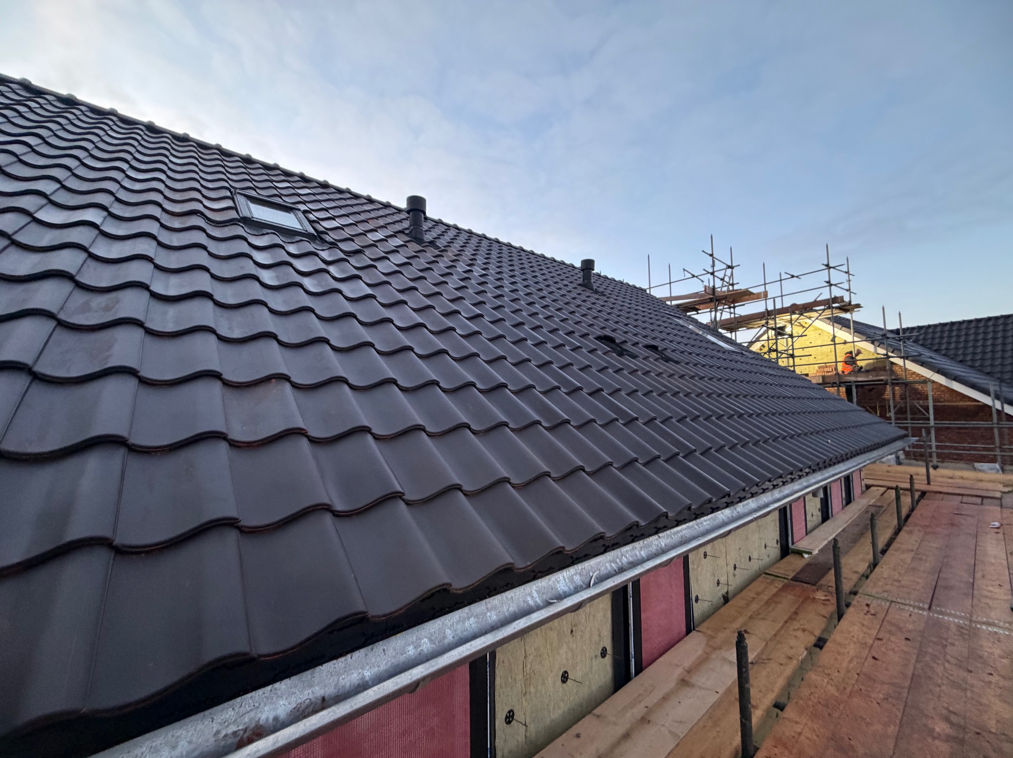 Ciemna dachówka ceramiczna na nowym dachu z oknem połaciowym i kominkami wentylacyjnymi. Widoczne rusztowania i ocieplenie poddasza.