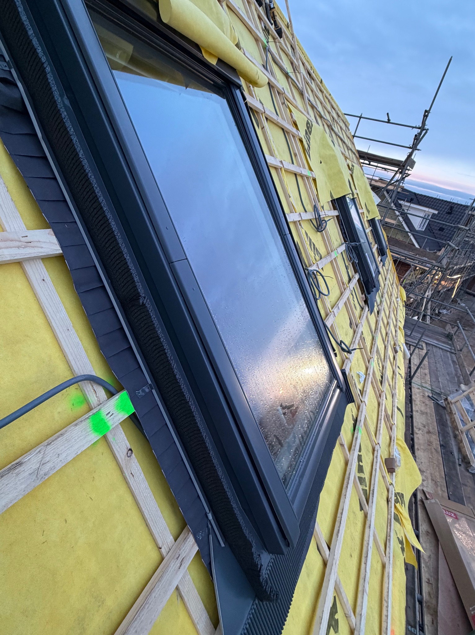 Montaż okna dachowego z widoczną membraną i łatami. Fragment dachu z szarą dachówką w trakcie prac dekarskich. Widoczne rusztowanie i fragment zabudowy w tle.