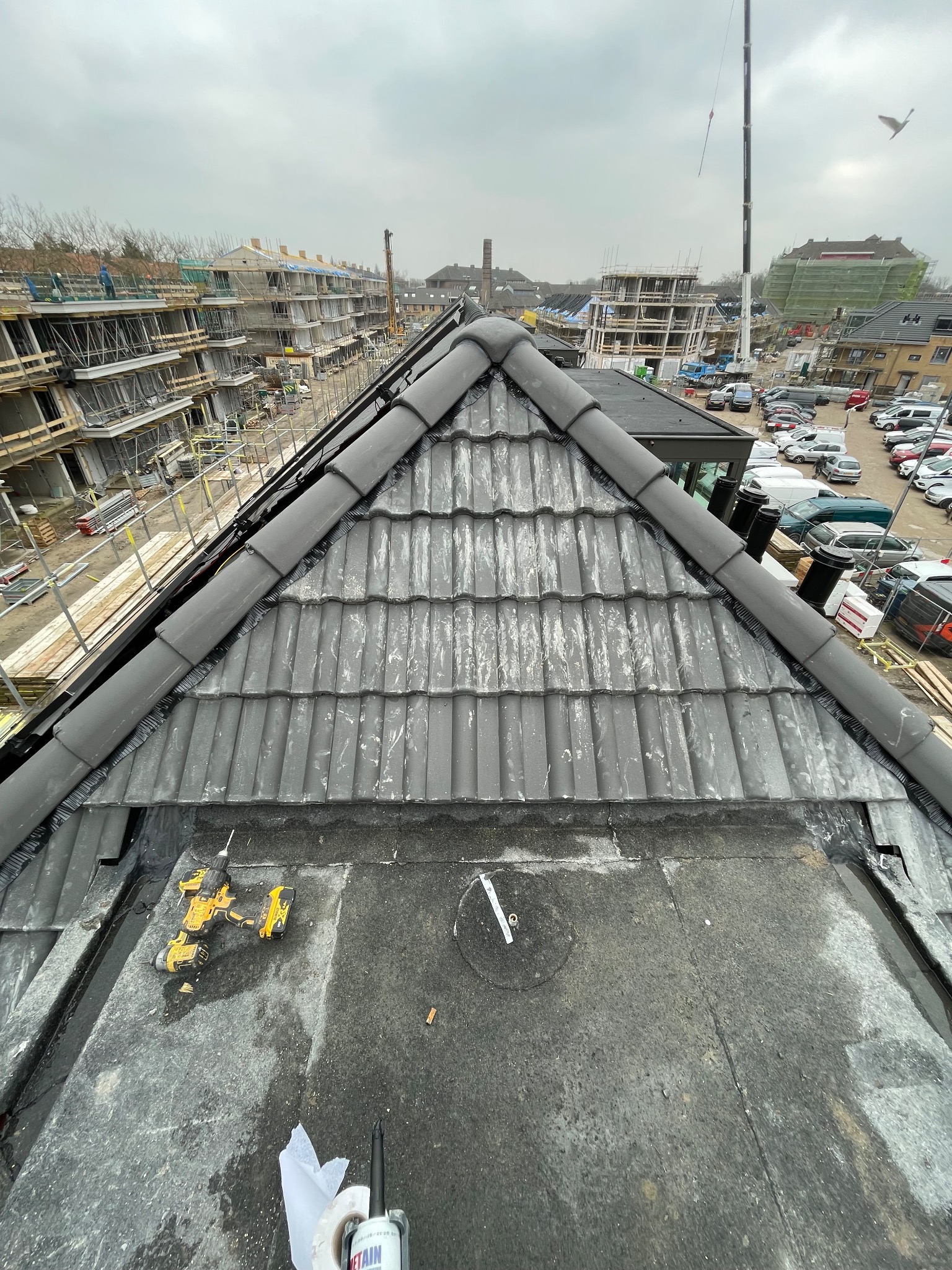 Szary dach z dachówki ceramicznej w trakcie montażu, widok z góry na narzędzia i fragmenty konstrukcji, w tle budowa nowych budynków i parking.