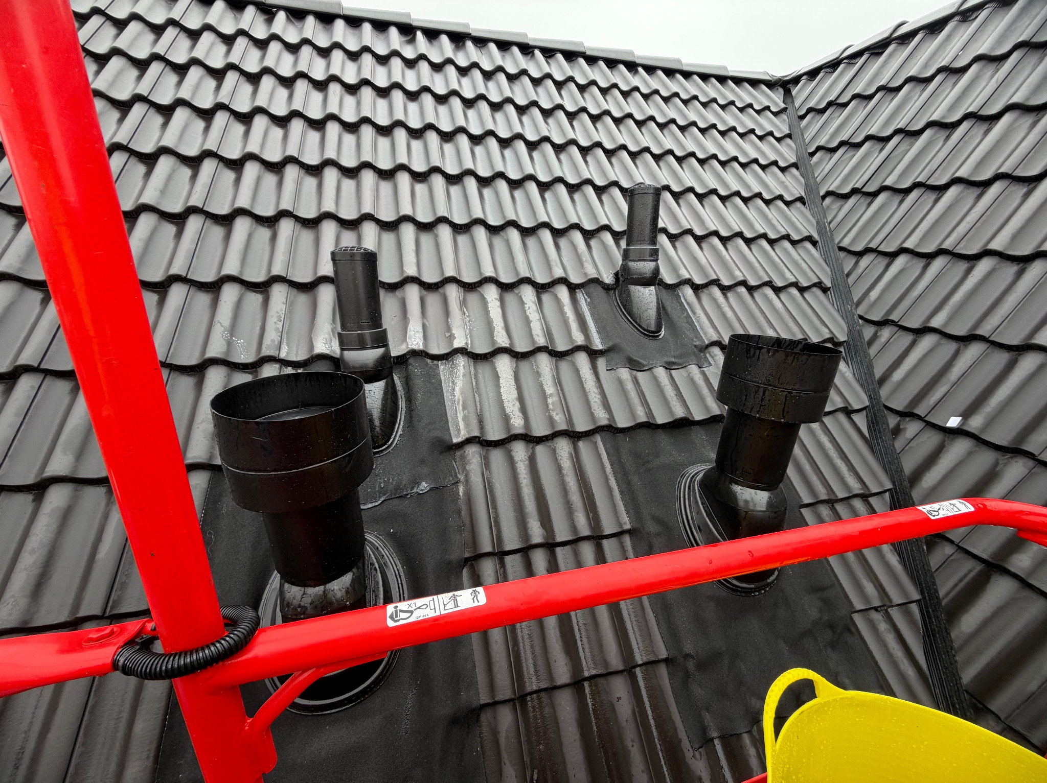 Czarny dach z dachówki ceramicznej z trzema kominami wentylacyjnymi, widziany z podnośnika koszowego z żółtym wiadrem. Detal roboczy.