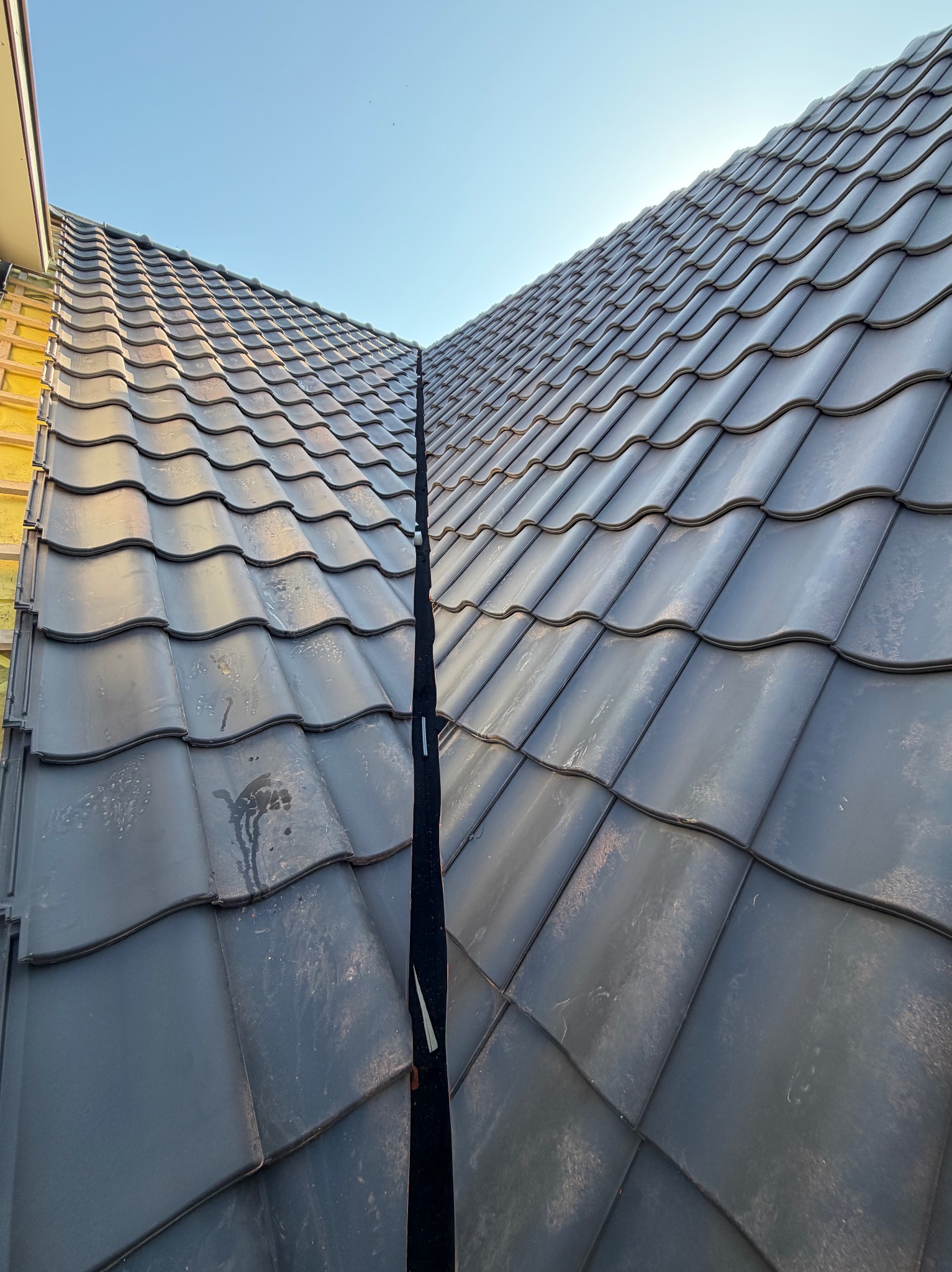 Nowy, ciemnoszary dach z dachówki ceramicznej, widok z góry, z wyraźnie zaznaczonym kalenicą i fragmentem ocieplonej ściany szczytowej budynku w tle.