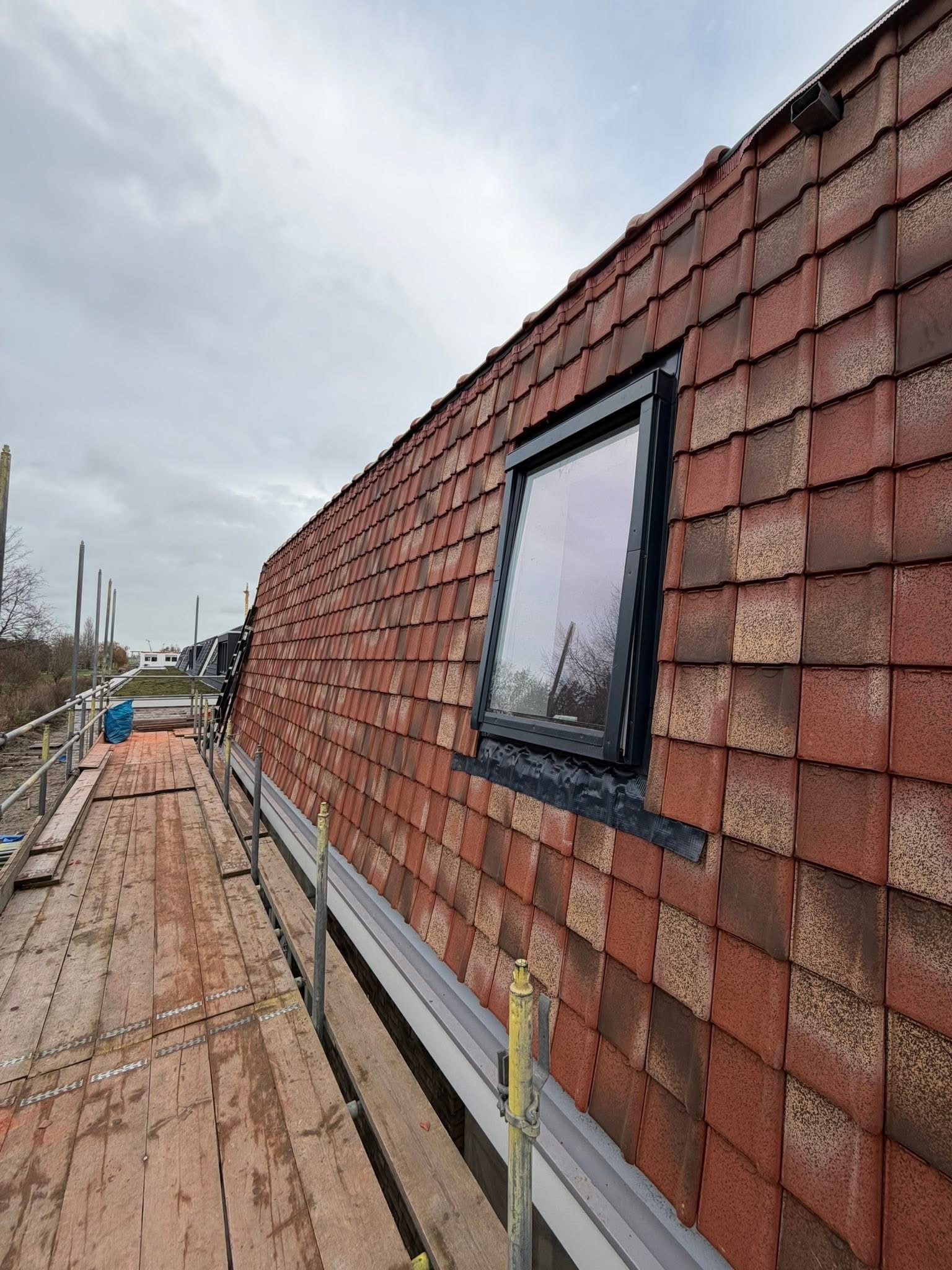 Czerwona dachówka ceramiczna z oknem dachowym w czarnej ramie, widok z rusztowania. Dach po renowacji, z bliska, w pochmurny dzień.