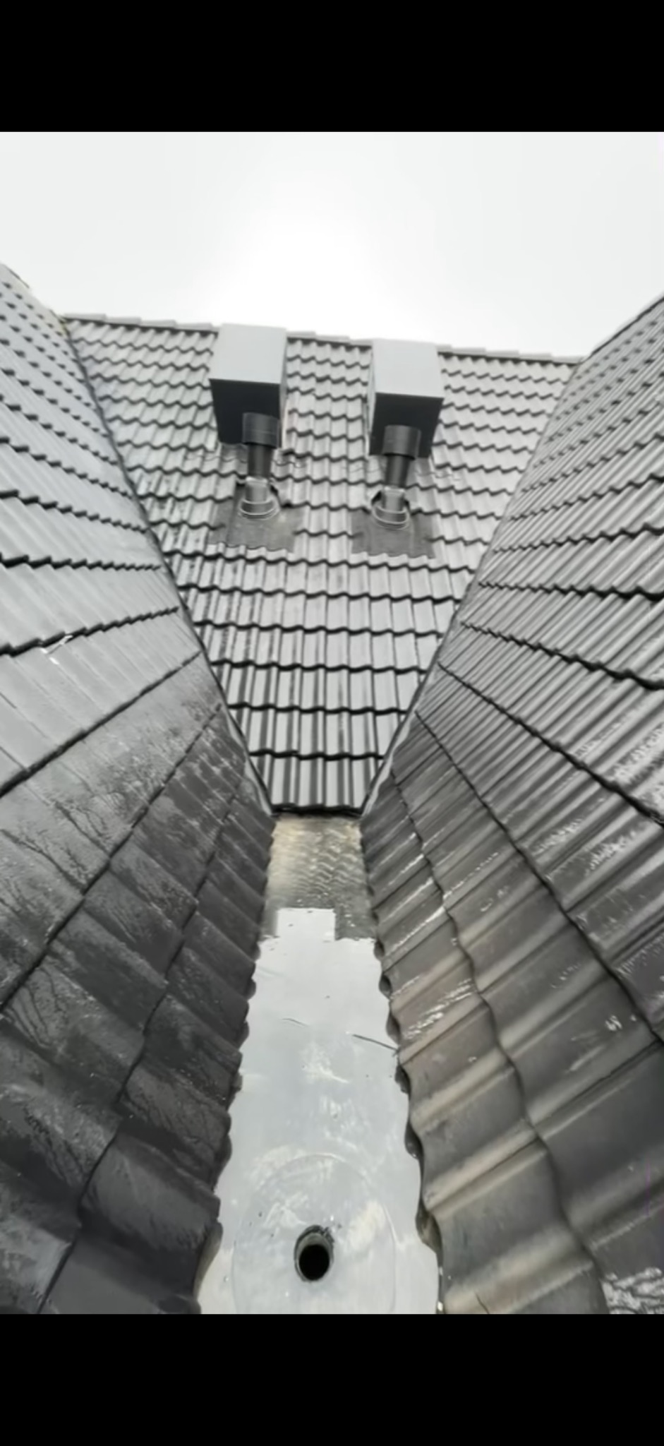 Czarny dach z dachówki ceramicznej po deszczu, widoczne dwa kominy wentylacyjne i woda zbierająca się w rynnie koszowej. Ujęcie z góry.