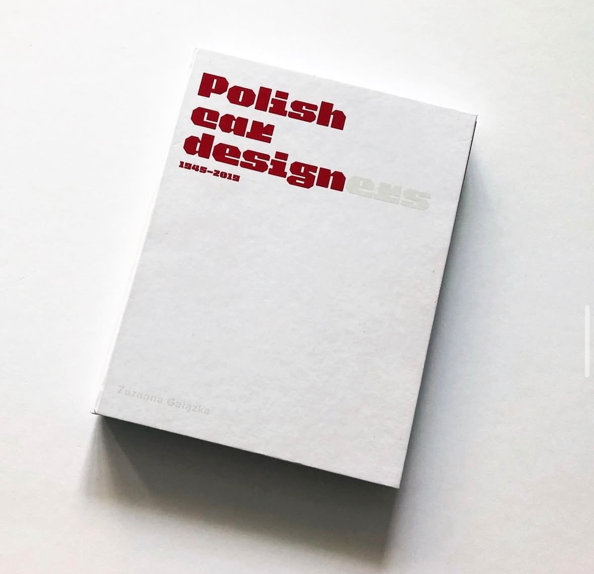 Książka 'Polish Car Designers 1945-2019' Zuzanny Gałązki na białym tle. Czerwony tytuł, minimalistyczny design okładki, widoczny cień.