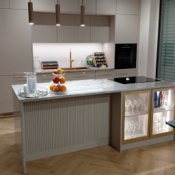 Stonekam - Beżowa kuchnia z wyspą i blatem z szarego kamienia. Na wyspie szklana patera z owocami. W tle zabudowa kuchenna z oświetleniem i złotą armaturą.