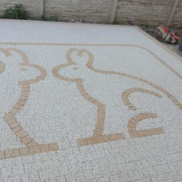 Stonekam - Ułożona kostka brukowa z dekoracyjnym wzorem dwóch zajączków w beżowym odcieniu. W tle widoczny fragment ogrodzenia i narzędzia.