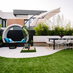 Stonekam - Nowoczesny taras z białych płyt, okrągła ławka z błękitnymi poduszkami, stół z krzesłami i parasolem. W tle bambusy i ogrodzenie.