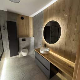 Bendix - Nowoczesna łazienka z drewnianymi i szarymi płytkami, okrągłym lustrem z podświetleniem LED, umywalką nablatową i szafką z szufladami. Minimalistyczny design.