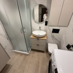 Bendix - Nowoczesna łazienka z kabiną prysznicową, okrągłym lustrem i umywalką nablatową. Beżowe płytki podłogowe i marmurowe na ścianach. Widoczna pralka i toaleta.