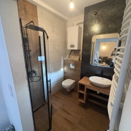 Bendix - Nowoczesna łazienka z kabiną prysznicową typu walk-in, drewnianą podłogą i szafką pod umywalkę. Na ścianie grzejnik drabinkowy i lustro z odbiciem remontu.