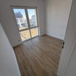 Bendix - Jasne panele podłogowe imitujące drewno w pustym pokoju z oknem, przez które widać budynki z panelami słonecznymi. Świeżo wykończone wnętrze.