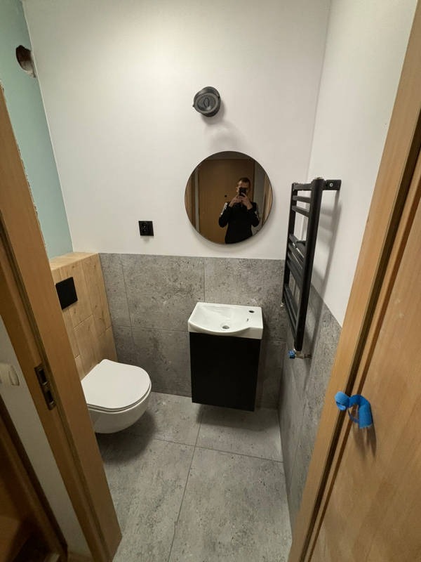 Nowoczesna, mała łazienka po remoncie: szare płytki, podwieszana toaleta i umywalka, okrągłe lustro z odbiciem osoby robiącej zdjęcie, grzejnik drabinkowy.