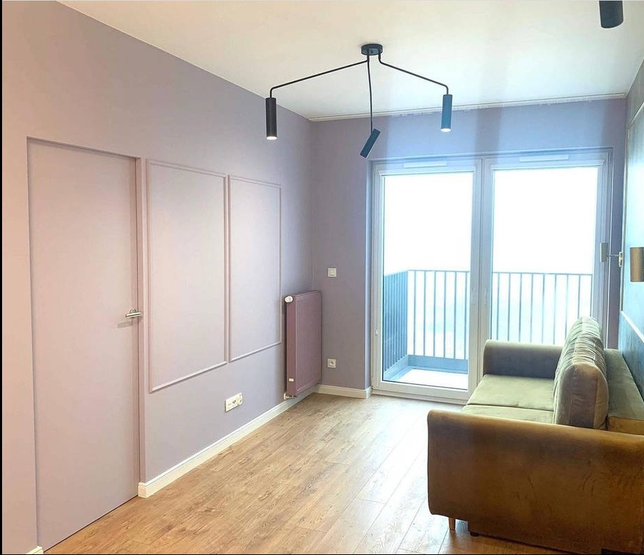 Pokój w pastelowych odcieniach z drewnianą podłogą, nowoczesnym oświetleniem i balkonem. Ściany zdobią dekoracyjne panele i dopasowany kolorystycznie grzejnik.